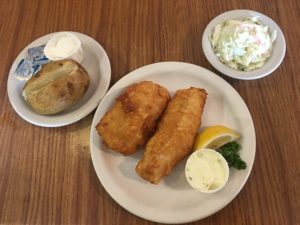 Fried fish sticks on a plate next to two smaller plates with a potato and coleslaw at Village Kitchen in Angola, IN