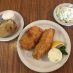 Fried fish sticks on a plate next to two smaller plates with a potato and coleslaw at Village Kitchen in Angola, IN
