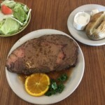 A large steak on a plate next to a slice of lemon and herbs with a potato and salad bowl at Village Kitchen in Angola, IN